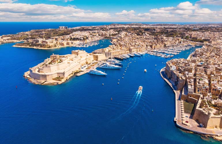 An aerial view of a port in Malta with the blue waters of the Mediterranean Sea highlighting the sandstone colored ancient fortress and buildings of the historic old city with superyachts docked along the coastline to illustrate the prestige of Malta's citizenship by investment program offered by Astons 