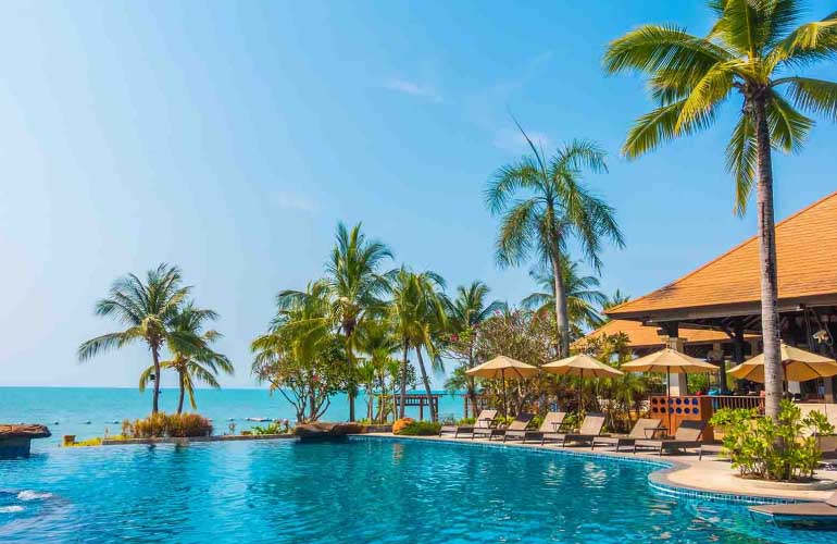 A picture of an exotic location showing a the large swimming pool at a luxury resort with palm trees and pool chairs next to the pool bar. The picture represents a typical luxury hotel in the Caribbean such as those that participate in citizenship by investment programs offered by Astons