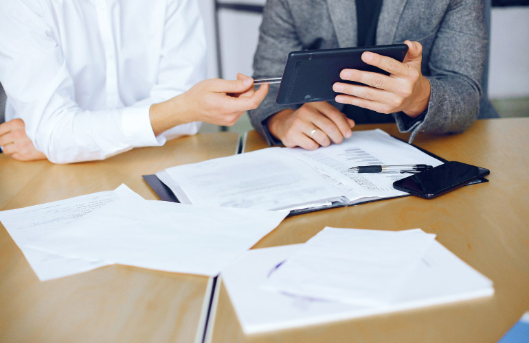 a few people working at a table, the table is covered in paperwork that looks like contracts, one person is holding a tablet explaining something to the other, the image depicts the legal process of due diligence as used in citizenship by investment, residency by investment (Golden Visas), and the luxury real estate transaction process