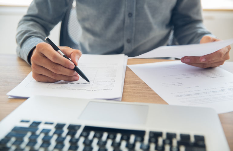 An image depicting a stage in the due diligence process common in the citizenship by investment or residency by investment process. A legal expert sit at his desk, with a laptop and paperwork in front of him, and he is carefully examining the contents of the paperwork
